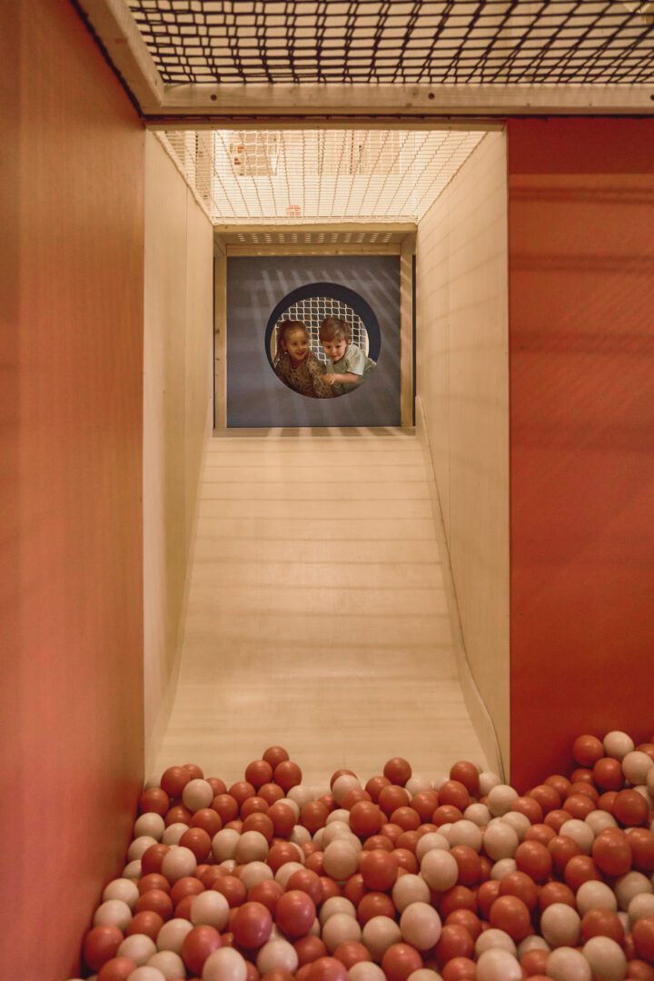 Dzieci wyglądają przez okrągłe przejście na pastelowy basen z kulkami o ciepłych kolorach. naturalny małpi gaj