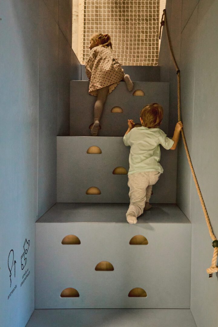 Dziewczynka i chłopiec wchodzą po schodach zabezpieczonych sznurkową poręczą i dziurkami do wspinania, rozwijając motorykę