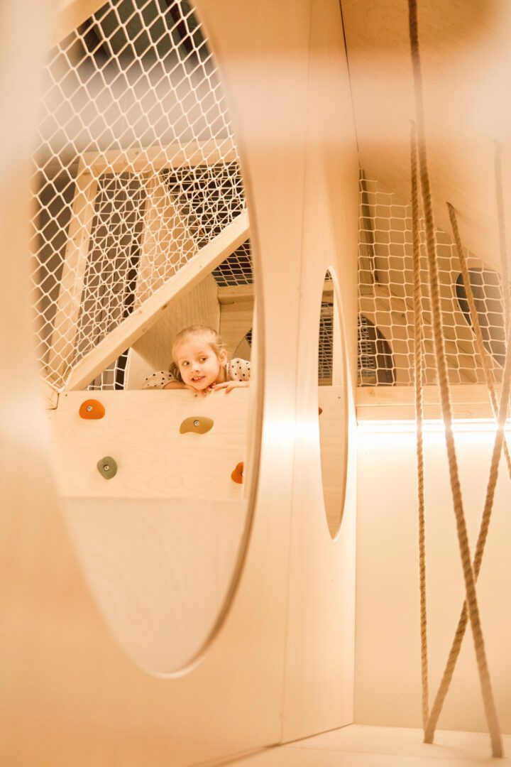 Dziewczynka wygląda przez otwarte przejście w drewnianym małpim gaju, wiele elementów so wspinaczki takich jak ścianka, czy siatka do wspinaczki