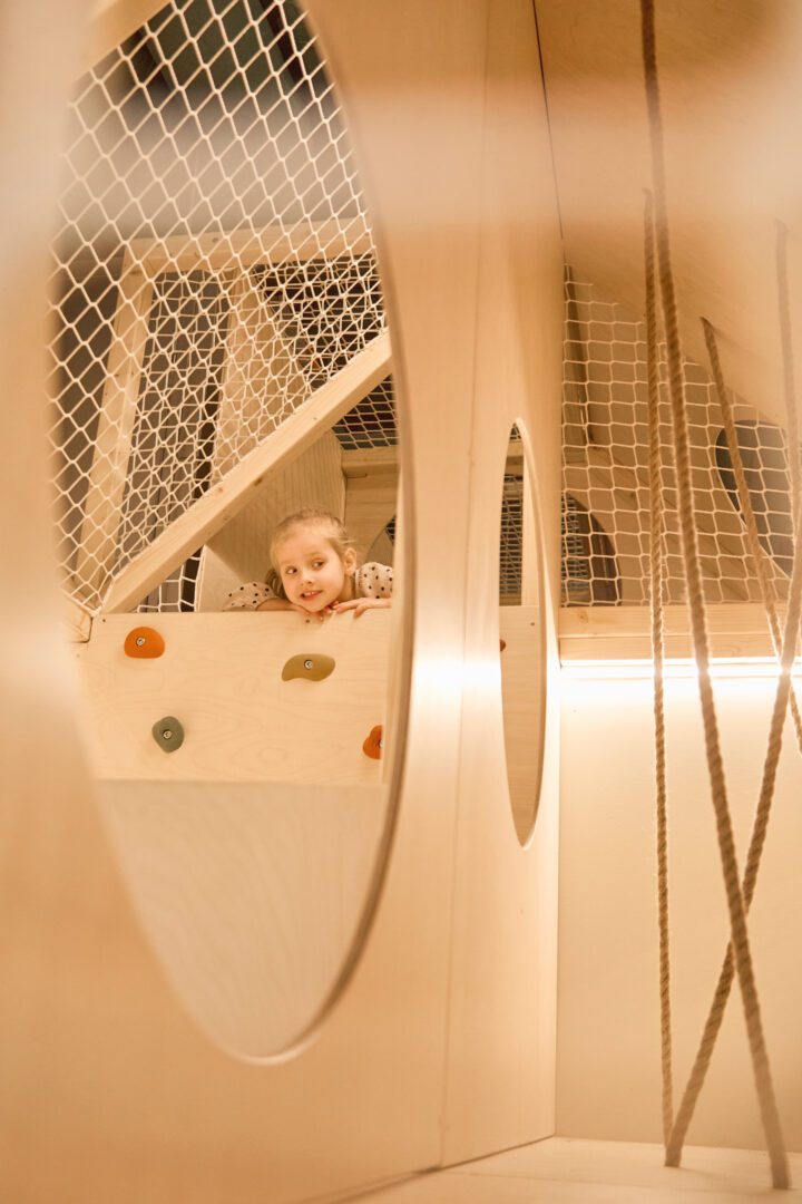 Dziewczynka po wspięciu na ściankę patrzy przez okrągłe przejście w drewnianym małpim gaju