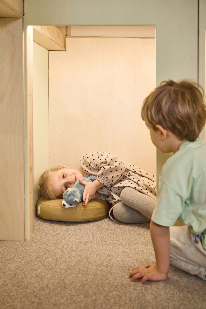 Dziewczynka leży na poduszce przytulając maskotkę, uśmiecha się, obok niej siedzi odwrócony do niej chłopiec